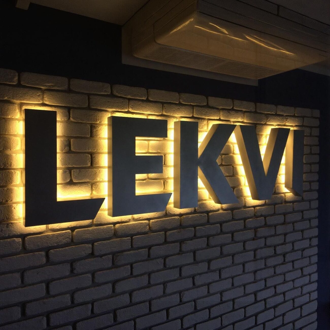 Czarne litery 3D z podświetleniem LED od tyłu, tworzące elegancki efekt HALO na ścianie z białej cegły.