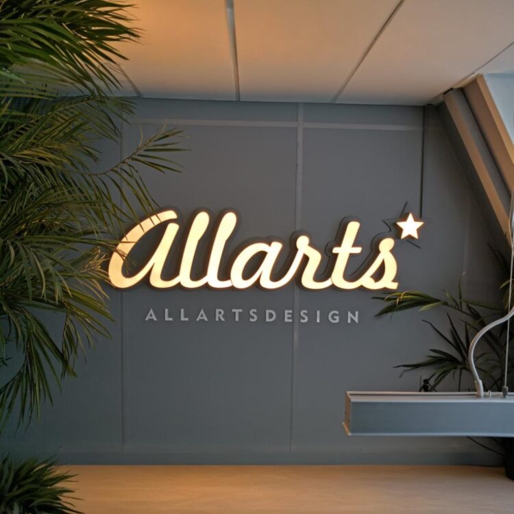 Podświetlany napis LEDON „Allarts” w ciepłym odcieniu światła, zamontowany na ścianie biura w otoczeniu zieleni.