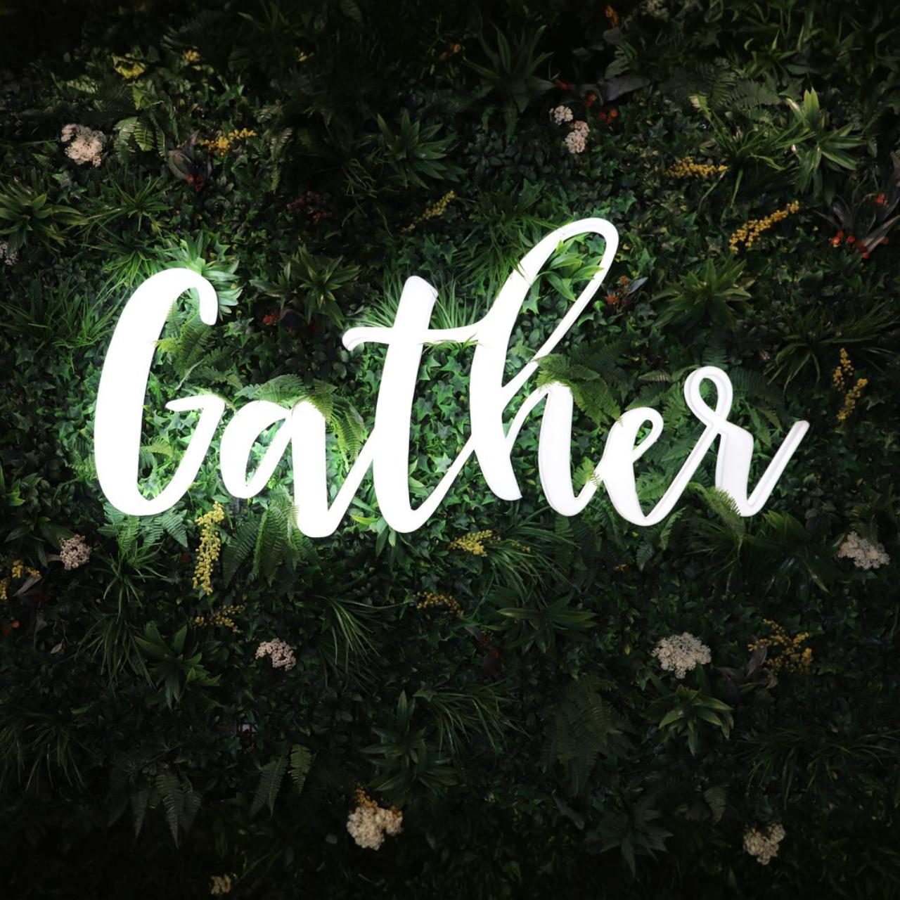 Podświetlany napis LEDON „Gather” zamontowany na tle ściany z roślin, wykonany z mlecznej plexi z subtelnym światłem LED.