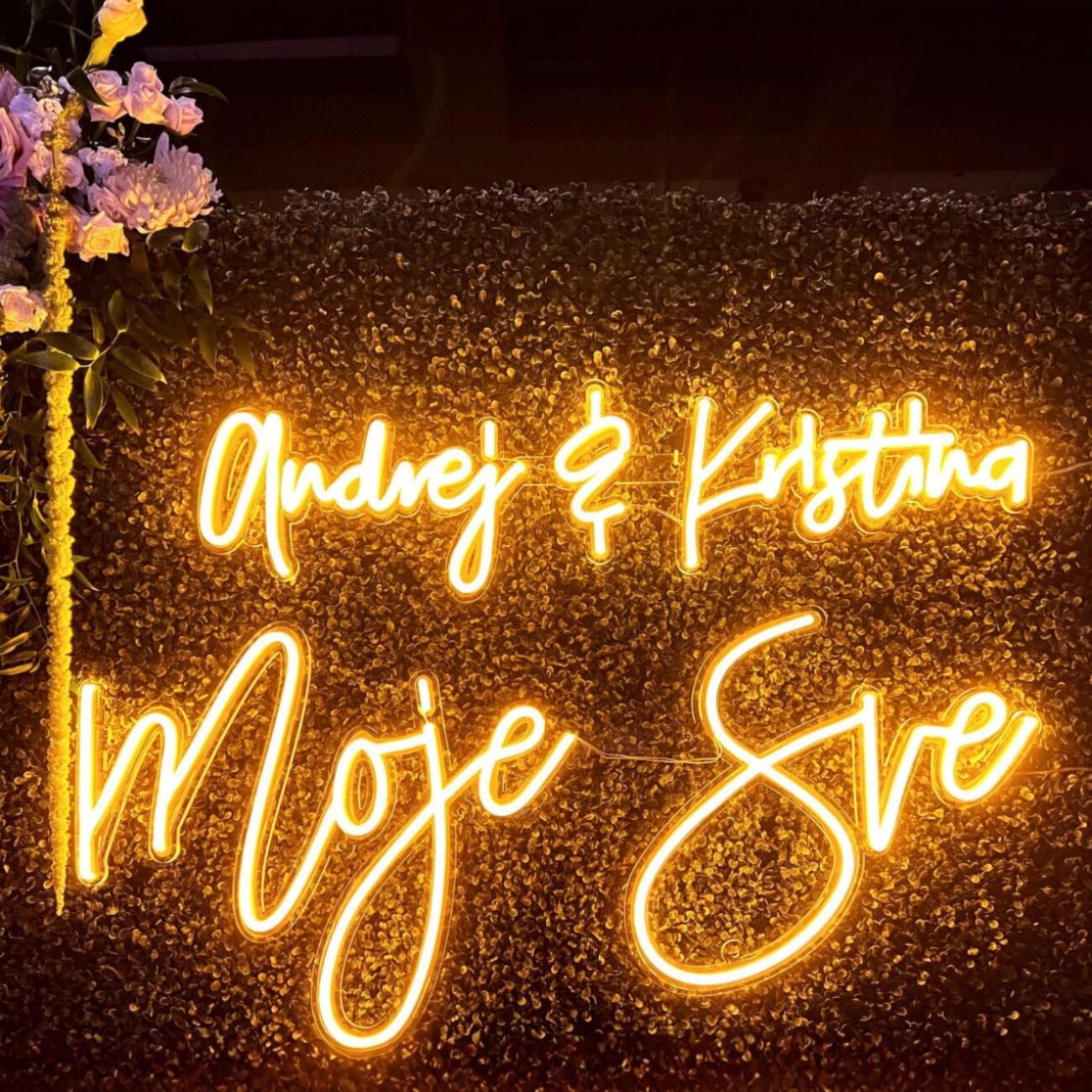 Neon LED o ciepłym, żółtym świetle z napisem dekoracyjnym, zamontowany na tle z zieleni i kwiatów – idealny na wesele lub event.