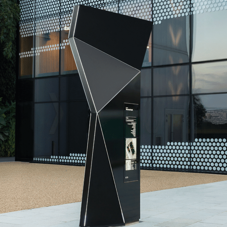 Czarny pylon informacyjny o geometrycznym kształcie z podświetleniem LED, stojący przed nowoczesnym budynkiem z przeszkloną fasadą.