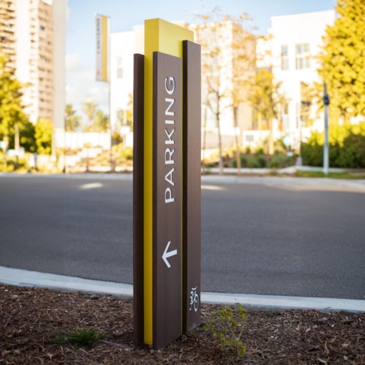 Nowoczesny pylon informacyjny z napisem „Parking”, wykonany z materiałów odpornych na warunki atmosferyczne, ustawiony przy wjeździe na teren budynku.