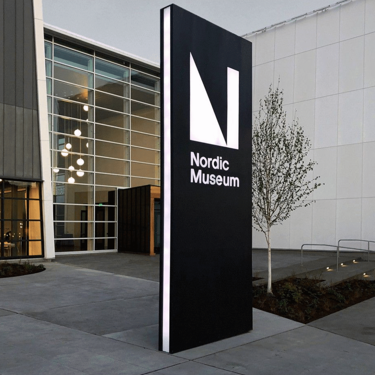 Wysoki pylon reklamowy z podświetleniem LED, ustawiony przy wejściu do nowoczesnego budynku z przeszkloną fasadą.