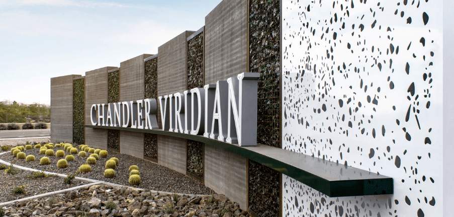 Nowoczesny pylon wejściowy z dużymi literami 3D umieszczony przed kompleksem budynków, otoczony zielenią i elementami architektury krajobrazu.