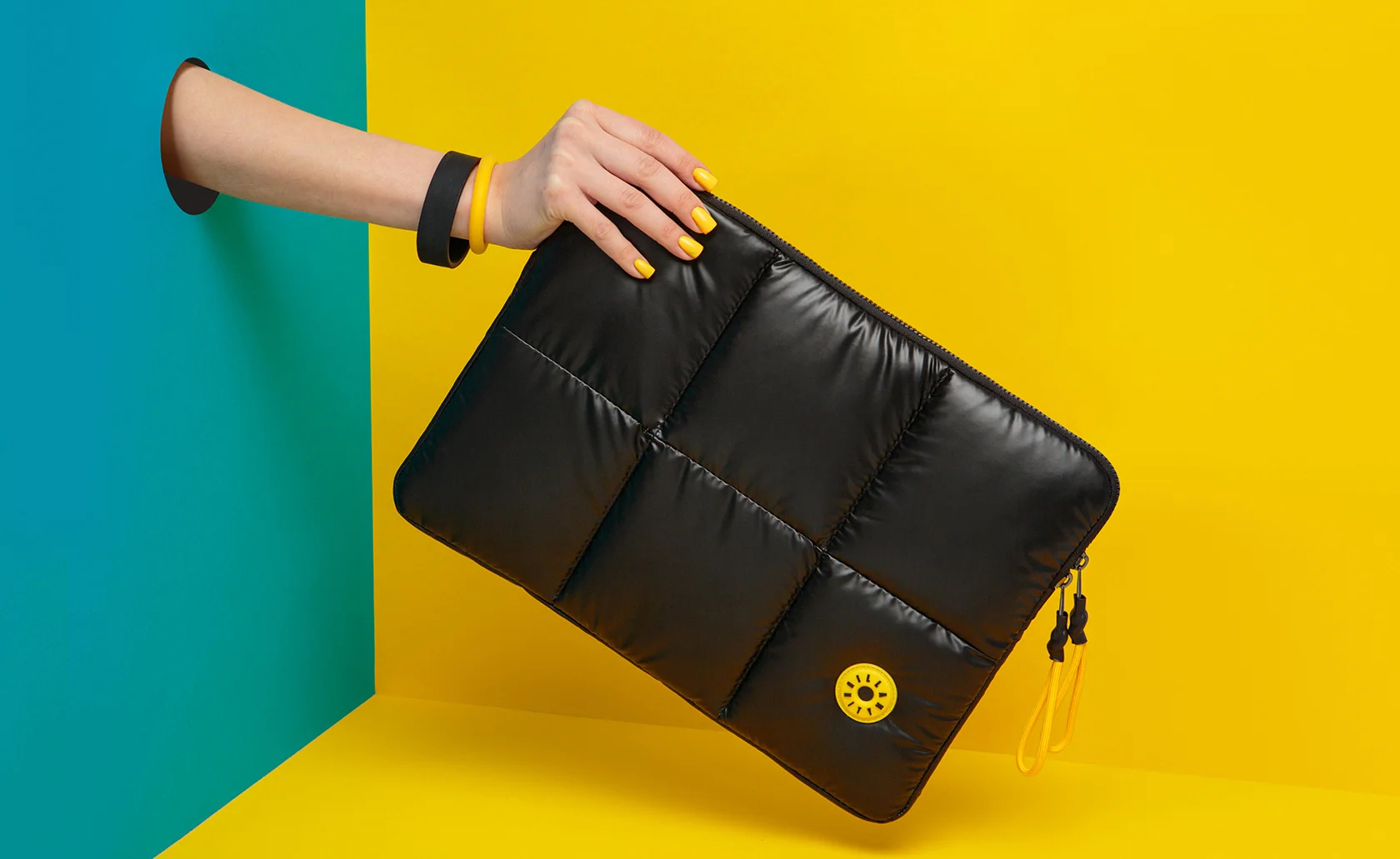 Ręka trzyma czarne etui z żółtym emblematem Illan Unity na tle turkusowo-żółtym.